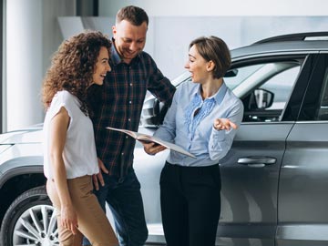 Ein Autoverkäufer bei Autoankauf Hamm bespricht Details mit einem Paar und hält dabei ein Broschüre in der Hand, in einem modernen Autohaus, mit Fahrzeugen im Hintergrund.